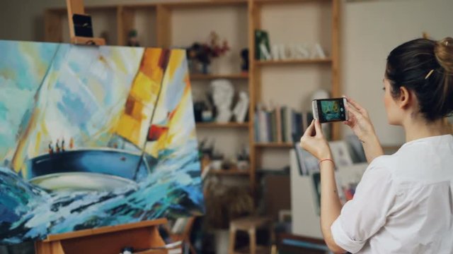 Female painter is taking photo of her beautiful picture using modern smartphone camera touching screen standing in studio alone. Modern technology and arts concept.