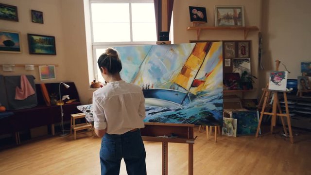 Pan shot of serious girl professional painter working in studio painting marine landscape with tempera paints holding palette and brush. Artworks and creativity concept.