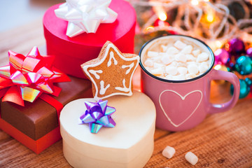 Cup of chocolate with marshmallow, gingerbread cookies, gifts and beautiful Christmas decorations with lights on the wooden background. Flat lay, top view, close up space for a text.