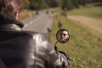 Smiling face of handsome bearded happy biker in dark sunglasses reflected in motorbike mirror on blurred background of sunny road and green rural landscape. Active lifestyle and love to adventures.
