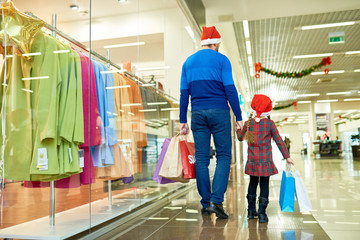 Back view of adult man and little girl in Santa hats walking in shopping mall buying Christmas gifts