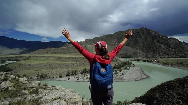 Young happy torist woman with backpack and cap standing on the edge of the mountain and puts his hands in the air. Back view, slider