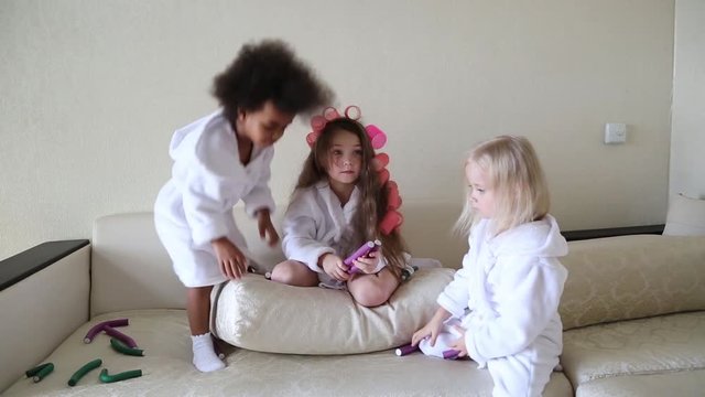 Little girls play with hair curlers and hairpins. Girls girlfriends in white coats braid curlers and hair clips each drg in her hair.