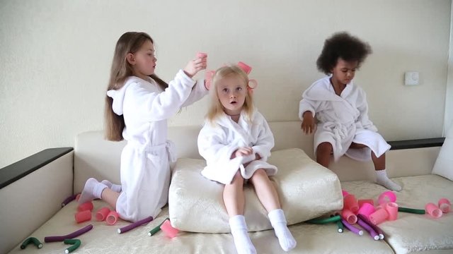 Little girls play with hair curlers and hairpins. Girls girlfriends in white coats braid curlers and hair clips each drg in her hair.