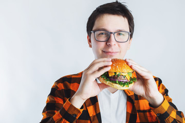 A young guy with glasses holding a fresh Burger. A very hungry student eats fast food. Hot helpful food. The concept of gluttony and unhealthy diet. With copy space for text