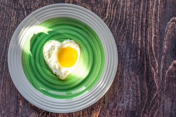 Fried egg in heart shape on a platter on wooden background. Top view.