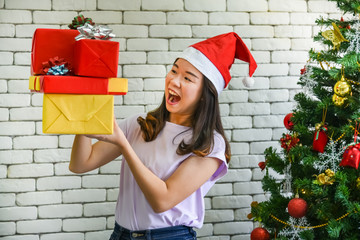 Asian girl happy smile, standing near the Christmas tree Decoration with the symbol of Christmas day, in her hand holds many gift box with white wall background, to New year and holidays concept.