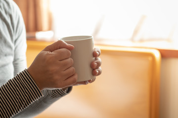 Close up of woman hand holding white mug cup of hot coffee with sunlight in the morning.