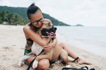Cheerful asian young woman in eyeglases sitting and hugging her dog on the beach