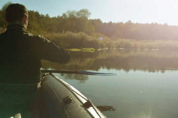 A man in black clothes and a cap swims in an inflatable boat with oars on the river on a clear autumn day in the rays of the sun.