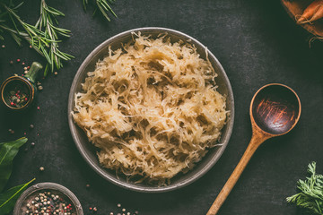 Pickled cabbage in bowl with wooden spoon and seasoning on dark rustic kitchen table background,...
