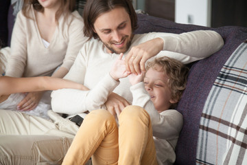 Obraz premium Young married couple with little preschool children playing sitting on couch in living room at new home. Parents and kids having a fun, close up. Happy family, buying real estate and mortgage concept