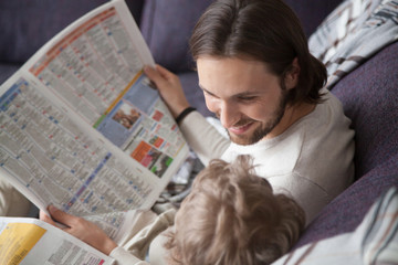 Close up family spending free time weekend at home. Father looking with love tenderness and laughing with little intelligent smart son reading news newspapers sitting together on couch in living room