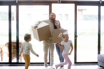 Married couple standing in living room with little children. Family moving at new house. Smiling parents holding unopened cardboard boxes, joyful preschool son and daughter running and playing around