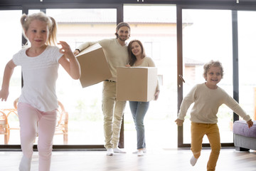 Fototapeta na wymiar Married millennial couple enter living room with little children. Family moving at new house. Smiling parents holding cardboard boxes, joyful toddler son and preschool daughter running playing around