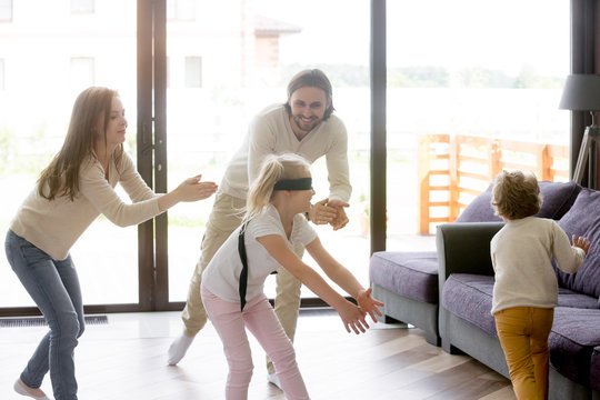 Happy funny family with little toddler son and daughter enjoy spending free time on weekend at new modern home. Playful small girl with covered eyes playing blindman game hide and seek in living room