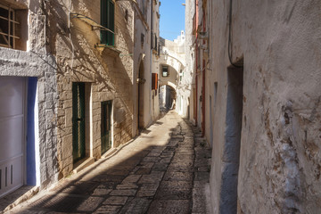 Fototapeta na wymiar A narrow street surrounded by vintage stone architecture and with an arch at the end in the old town area of the town of Ostuni in the Puglia region of Italy on a summer day with a clear blue sky