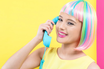 Young woman in a colorful wig talking on a retro phone on a split yellow and pink background