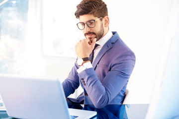 Thinking young businessman sitting behind his laptop while working in the office
