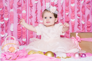 Obraz na płótnie Canvas Cute little baby girl sits on a soft blanket in a pink dress with a Christmas balls, party gift and handbag, in a festive pink interior. New Year and Christmas holidays.