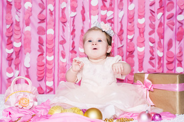 Obraz na płótnie Canvas Cute little baby girl sits on a soft blanket in a pink dress with a Christmas balls, party gift and handbag, in a festive pink interior. New Year and Christmas holidays.