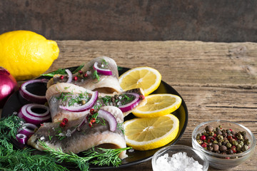 herring fillet with pepper, herbs, onion and lemon on black plate on rustic wooden background. close up