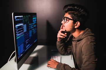 Thoughtful young indian programmer coding on computer in the evening at home
