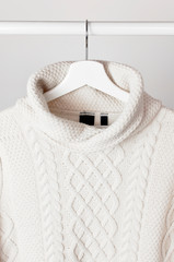 White knitted wool sweater on a white wooden hanger against the background of the light wall....