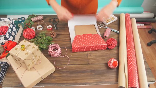 Female hands wrapping Christmas gift in craft ecological paper on dark wooden backround with decorating elements, colourfil twine stripes and pack paper rolls, Christmas or New year DIY packing.
