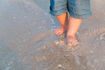 Bare feet walking at sandy beach near the sea. Little baby in blue jeans shorts going to touch the sea at sunset. Wave washes baby's feet. Toned. Soft focus.