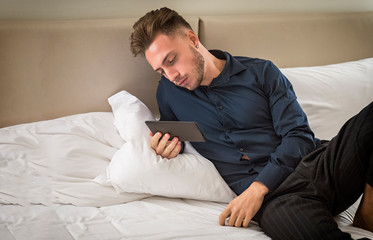 Fototapeta premium Handsome young man at home reading with ebook reader lying on his bed