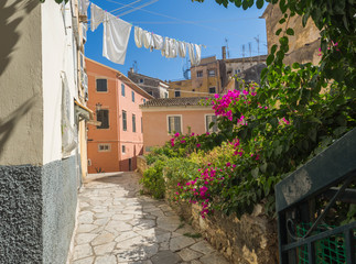 Corfu old town narrow cobble stone street with pink Bougainvillea flowers, traditional greek houses with hanging drying white laundry, blue sky summer sunny day