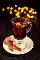 closeup glass of mulled wine with orange and cinnamon on dark black background, on white plate, Christmas lights