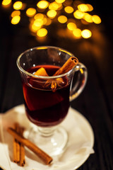 closeup glass of mulled wine with orange and cinnamon on dark black background, on white plate, Christmas lights, large yellow bokeh