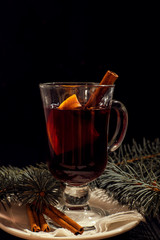 closeup glass of mulled wine with orange and cinnamon on dark black background, on white plate, christmas ball, tree