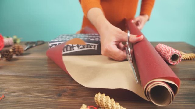 Overhead shot of female designer hands packing Christmas presents in wrapping papers on the wooden table with decorating items and Christmas baubles