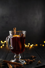 closeup glass of mulled wine with orange and cinnamon on dark black background,  Christmas tree and lights, large yellow bokeh, mulled wine set
