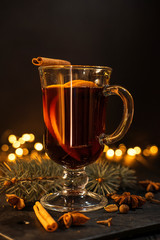 cinnamon lies in a glass, closeup glass of mulled wine with orange and cinnamon on dark black background,  Christmas tree and lights, large yellow bokeh, mulled wine set
