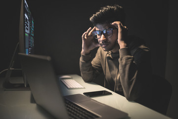 Concentrated sad asian young man in glasses sitting at the table and using computer in dark room