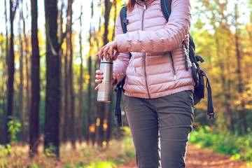 Refreshment in forest during autumn hiking. Female traveler with backpack holding a thermos