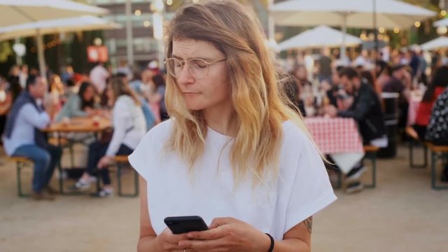 Young blonde girl or millennial hipster uses smartphone navigation or messaging application to meet with friends or family, searches for someone in crowd. Concept technology and everyday life
