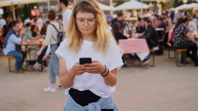 Confused and serious young woman or millennial hipster texts friends to meet up in crowd of party festival, can't find someone, waves and smiles. concept relationship or get together