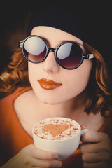 portrait of a beautiful girl with curly hair dressed in a beret and sunglasses holding a cup of coffee against a red background.