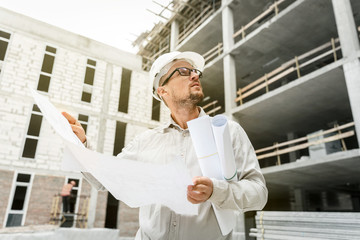 Construction engineer in a white hardhat inspecting blueprints on a construction site.