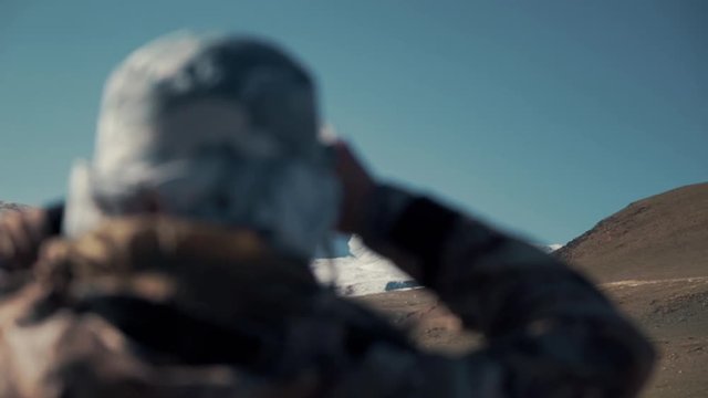 Scientist is watching wild mountain sheep (argali) through his binoculars. Snow peak and mountains in the background. Back view
