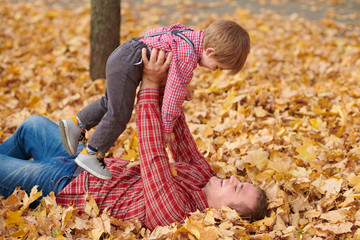 Father and son are lying on yellow leaves and having fun in autumn city park. They posing, smiling, playing. Bright yellow trees.