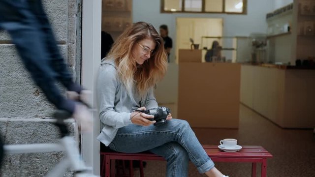 Cinematic shot of casual beautiful young blonde woman tourist, traveler or freelance creative photographer look through photos on professional camera at cafe on busy street. Urban nomad