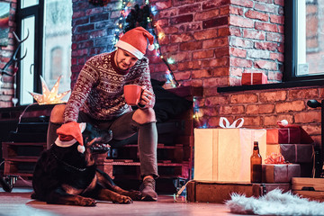 Obraz premium A young man holding cup with coffee during Christmas time with his cute dog in a decorated living room.