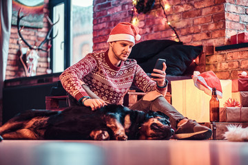 A young stylish man holding a smartphone while sitting with his cute dog in a decorated living room at Christmas time.