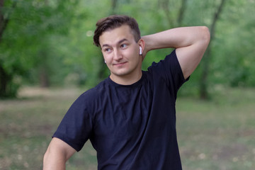 Young handsome caucasian man puts a hand on the neck, complicated expression on the face, smiling. Outdoors, green park background, copy space, close up. Casual wear, white earphones, trendy.